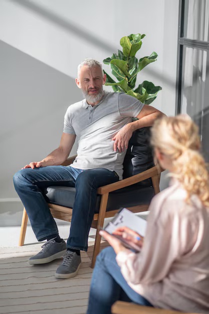 Hombre mayor sentado en un sillón conversando con una mujer que toma notas en una libreta, en un entorno luminoso y moderno. Una planta decorativa se encuentra al fondo.